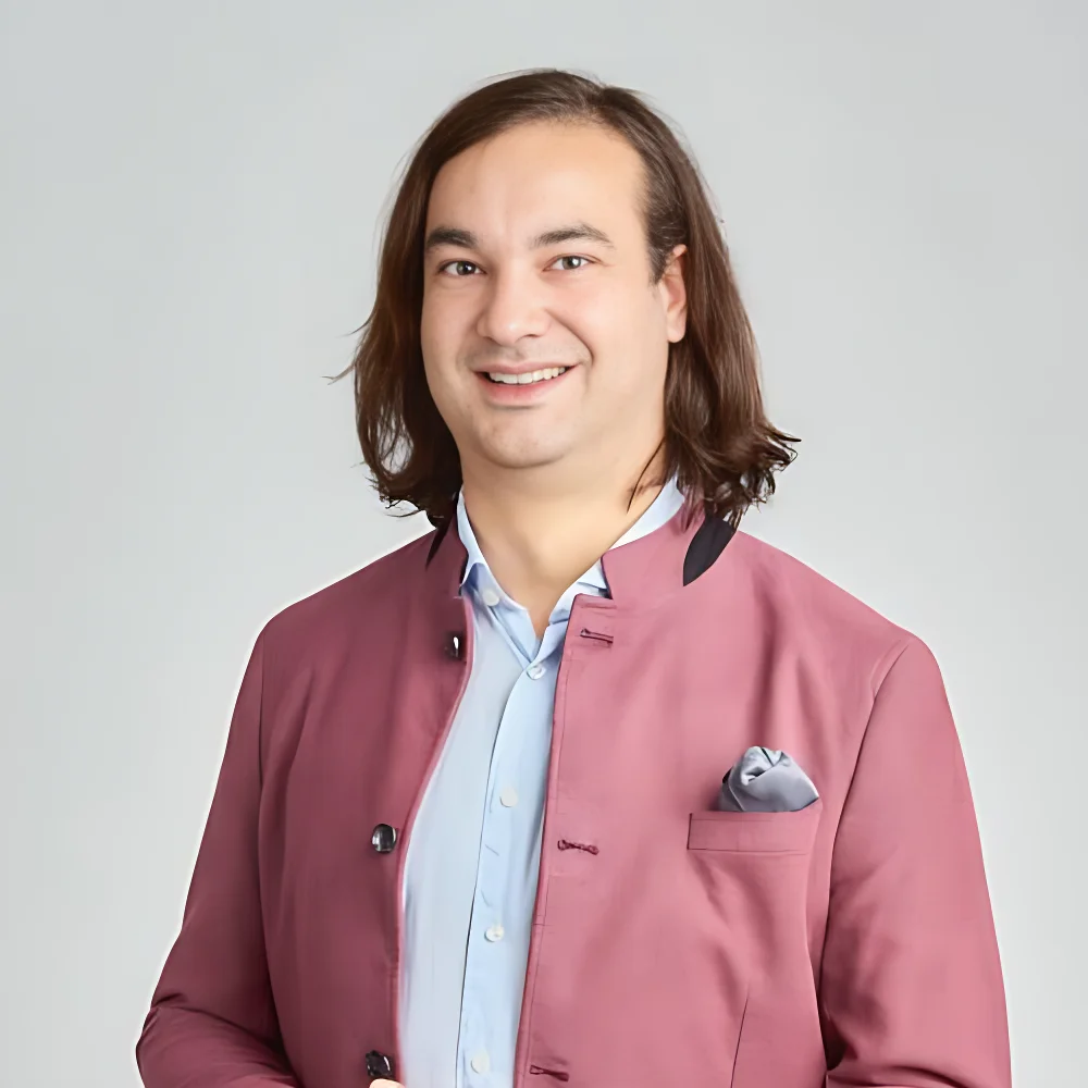 Porträt Von Michael Kaulartz, Standortleiter Von Home Music Teachers Mit Mittellangen Braunen Haaren, Hellblauem Hemd Und Bordeauxrotem Sakko, Vor Neutralem Hintergrund.